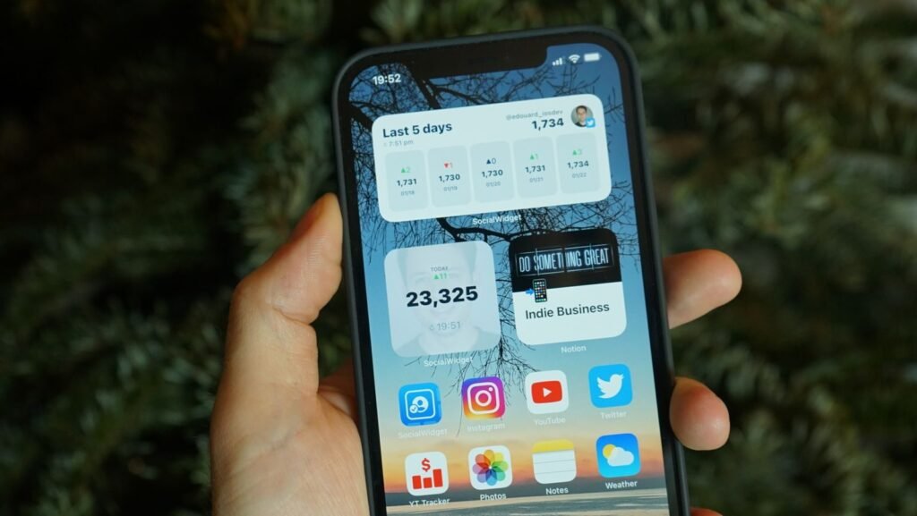 A hand holding a smartphone displaying various home screen widgets and app icons.
